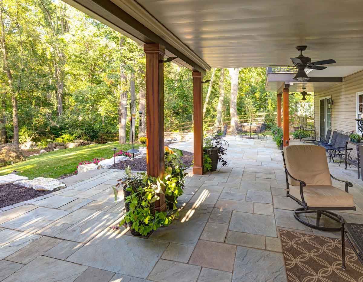 Covered patio with stone floor, seating, and lush greenery.