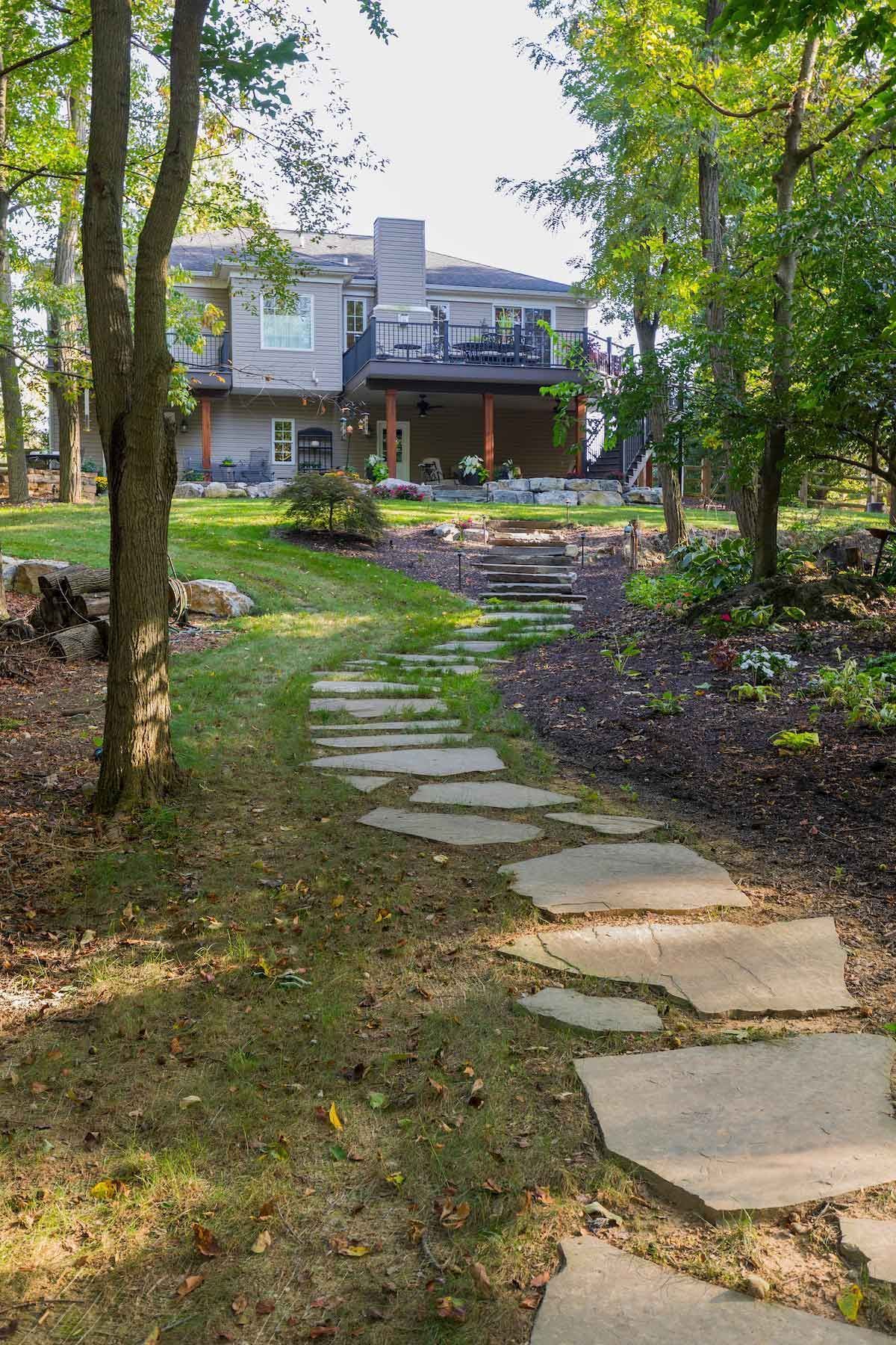 Stone path leads uphill to a house with a deck, surrounded by trees and lawn.