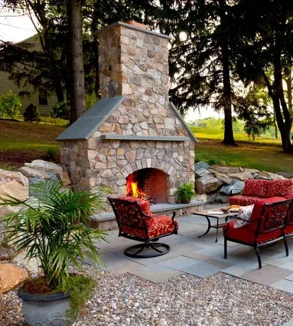Outdoor stone fireplace with seating area on a patio. Red cushions, green trees, and a sunny backyard.