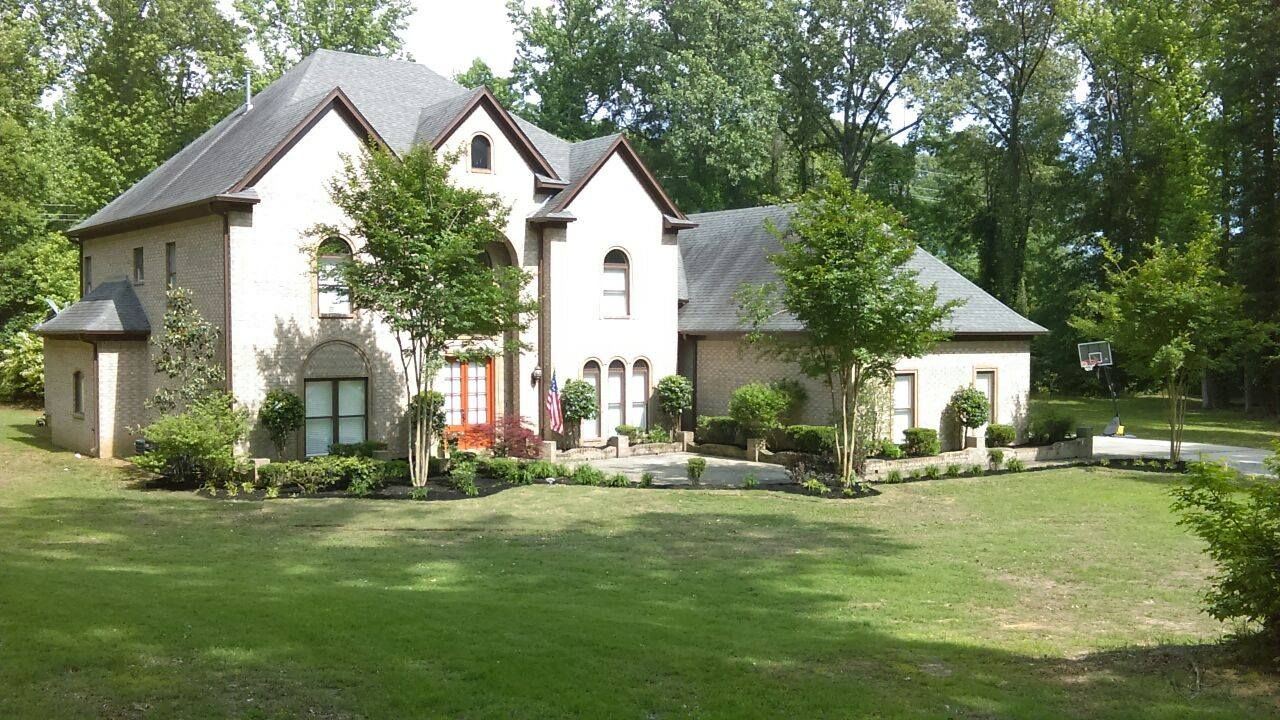 Two-story beige brick house with black roof, green lawn, trees, and small shrubs.