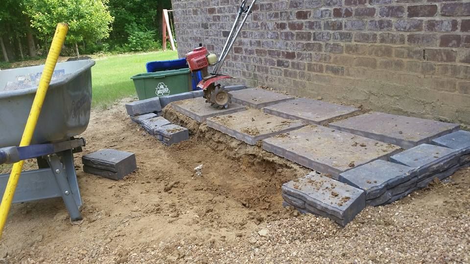 Construction site with pavers, sand, retaining wall, and a brick building in the background.