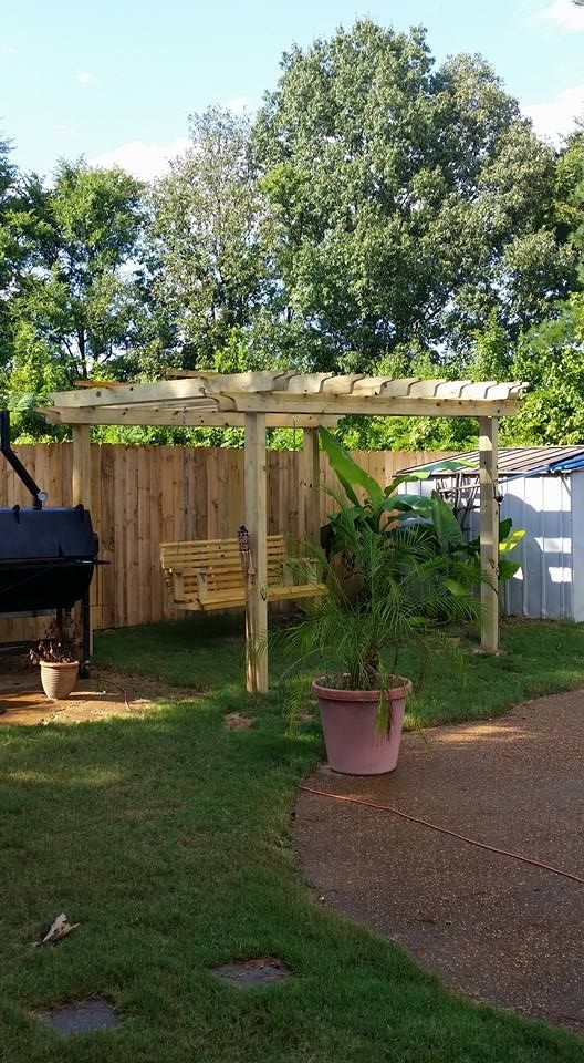 Wooden pergola with swing, surrounded by a wooden fence, grill, potted plants, and trees.