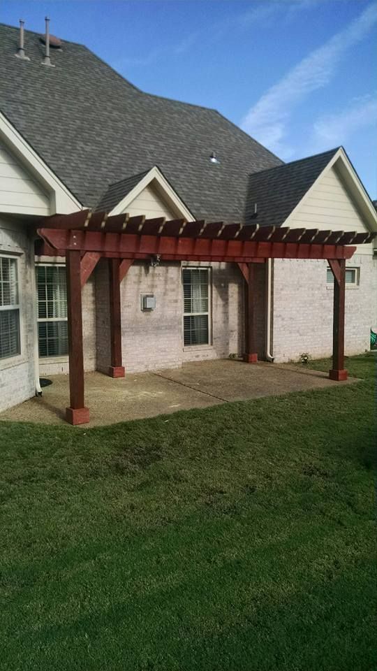 Red pergola attached to a brick house over a concrete patio, with green grass in the foreground.