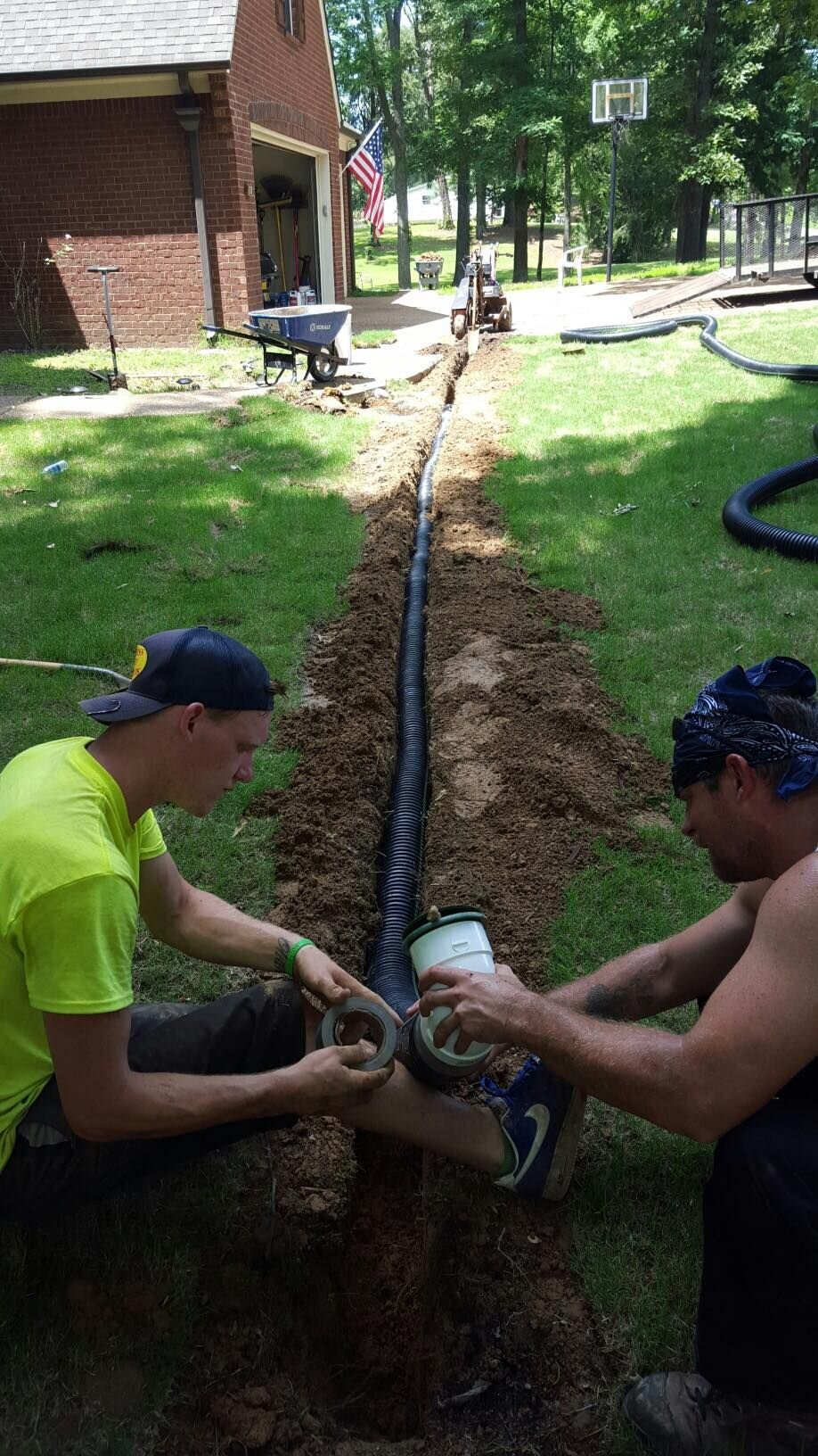 Two people installing a black drainage pipe in a trench.
