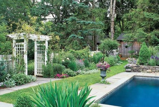Garden with a white pergola, green grass, and a blue-tiled pool. Lush plants surround the space.