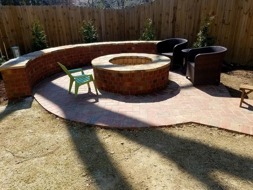 Brick patio with built-in fire pit and curved seating. Two chairs, one green, one brown. Wooden fence in background.