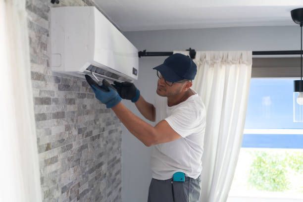 Man in cap and gloves inspecting a white wall-mounted air conditioning unit on a brick wall.