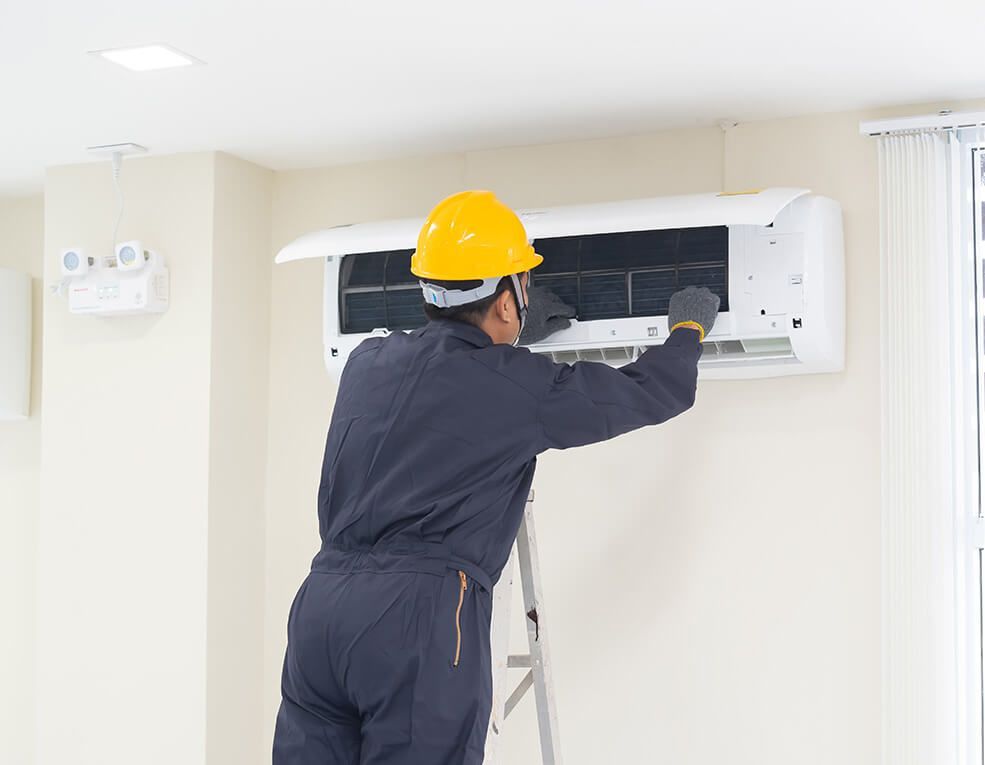 Person in blue jumpsuit and yellow hardhat services a wall-mounted air conditioner, using a ladder.