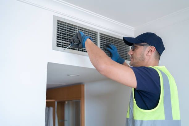 HVAC worker in a reflective vest cleans a vent. Indoors, white walls, blue gloves, cap.