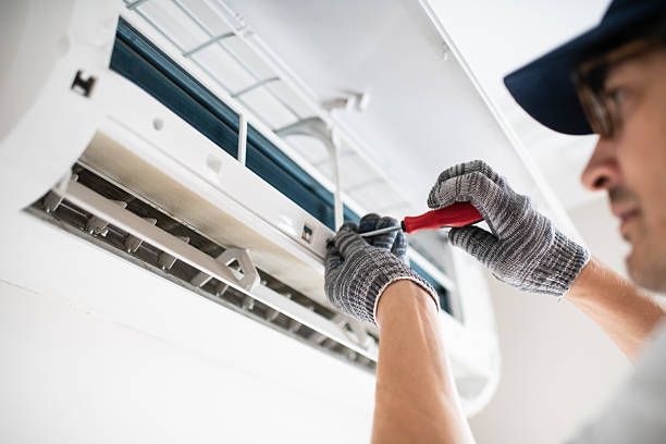 A person in a cap and gloves is repairing a white air conditioner unit with a screwdriver.