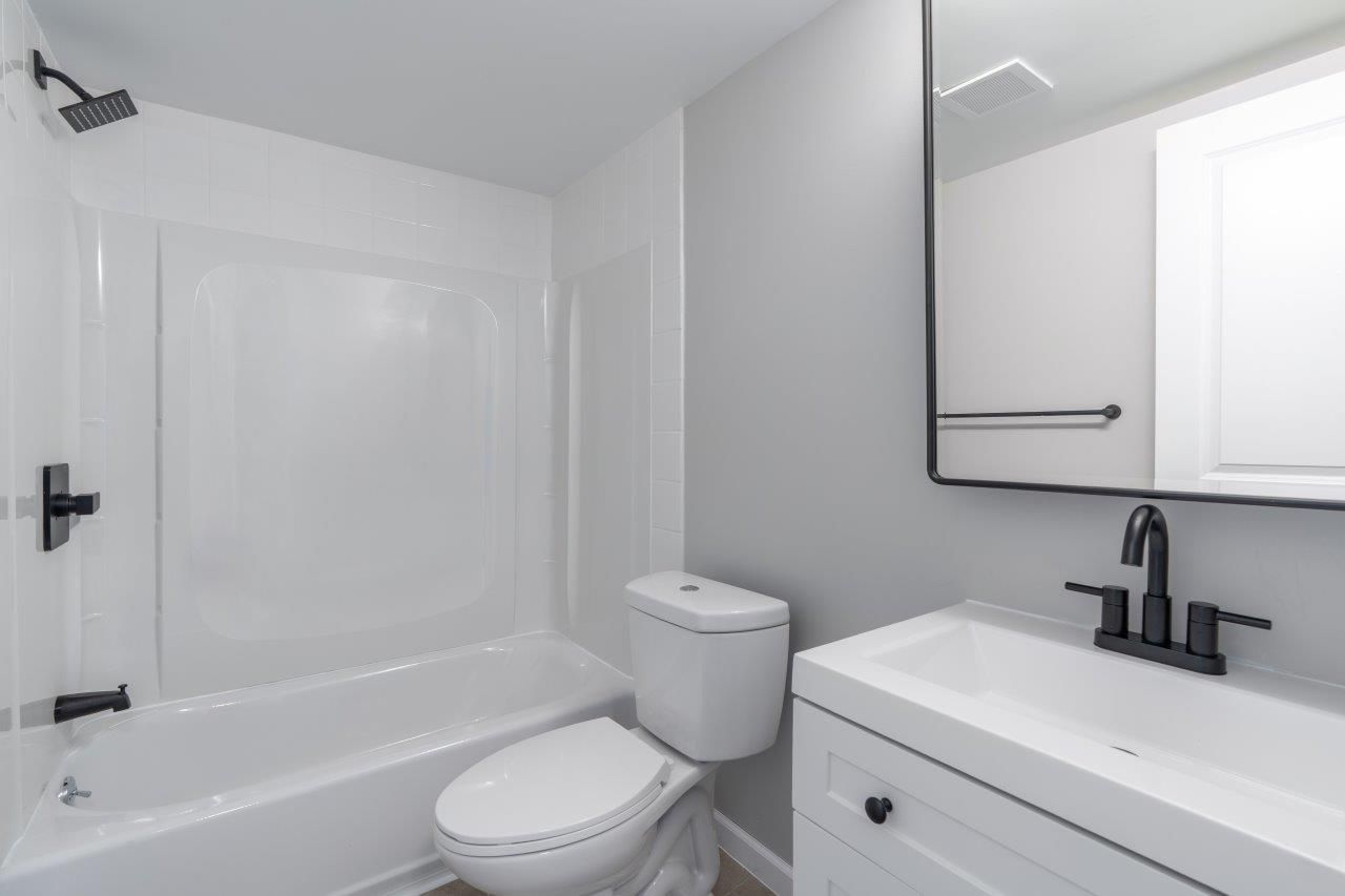 White bathroom with bathtub/shower, toilet, and vanity with black faucet.