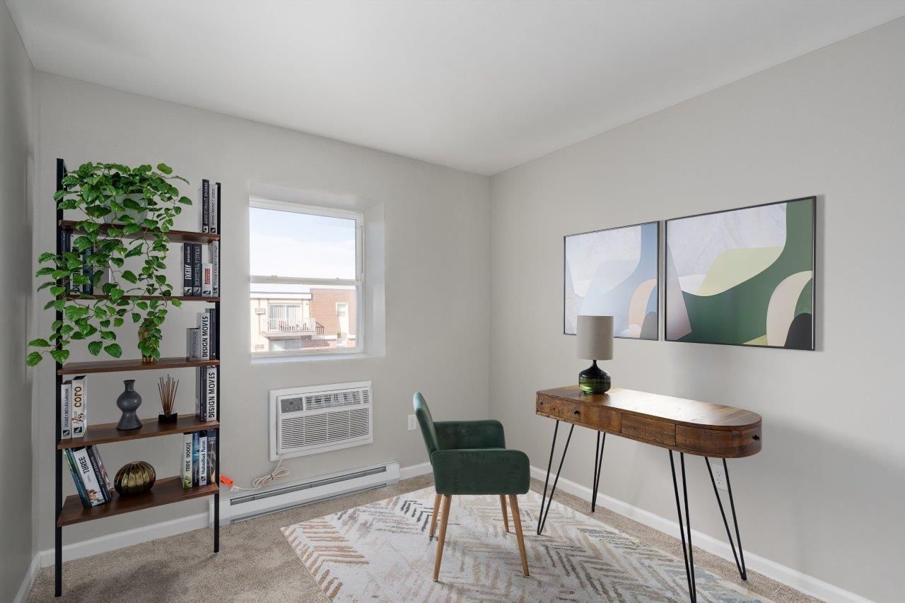 Apartment home office with desk, green chair, bookshelf, and plant near a window.