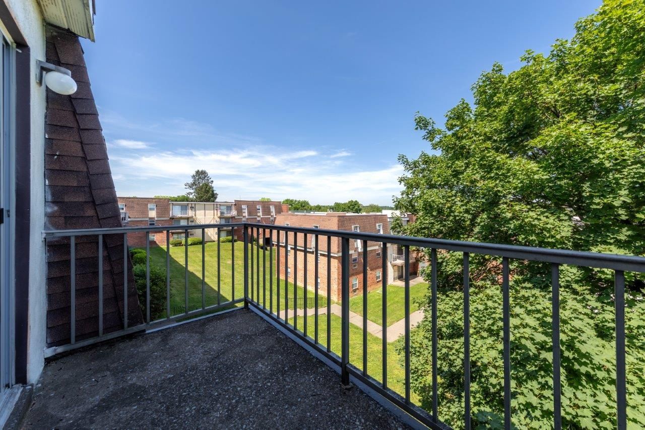 Balcony with black railing overlooking green trees and brick apartment buildings under a clear blue sky