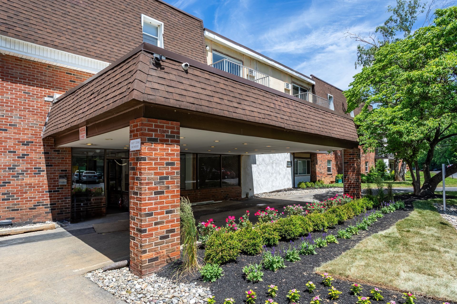 Brick apartment building entrance with covered walkway and landscaped flower bed under a blue sky