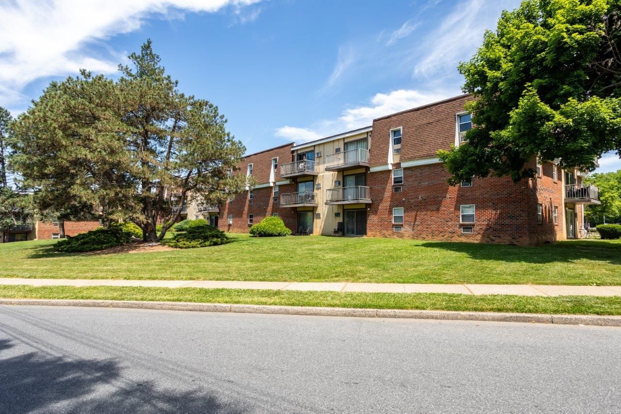 Brick apartment building with balconies, green lawn, and trees beside a quiet street
