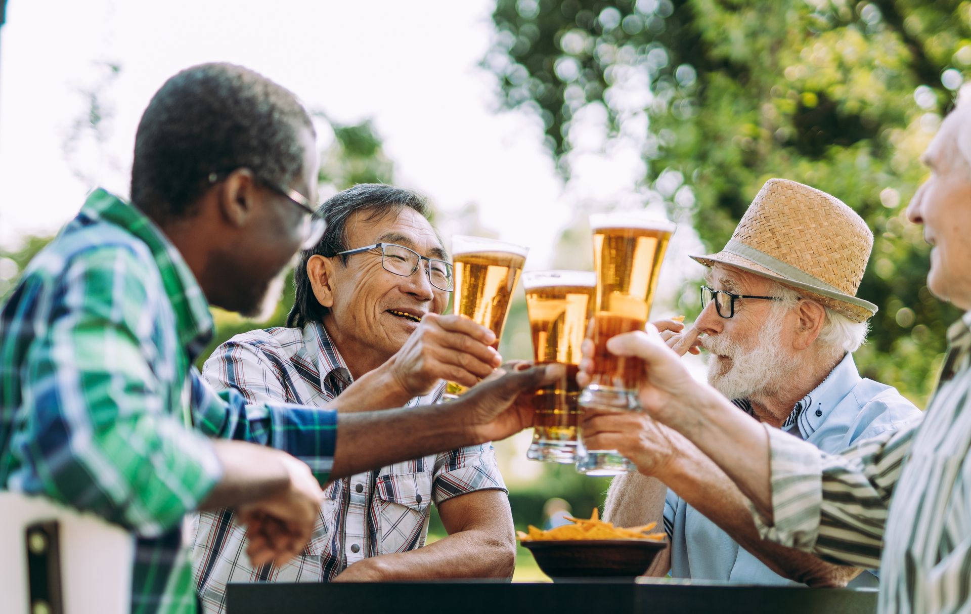 Four diverse senior men clink beer glasses outside, smiling and celebrating.