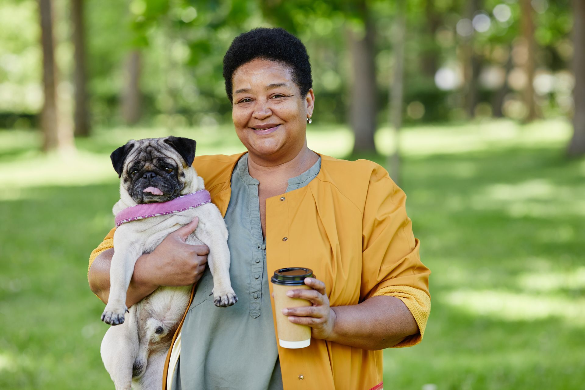 Woman in park holding pug dog and coffee, smiles at camera.