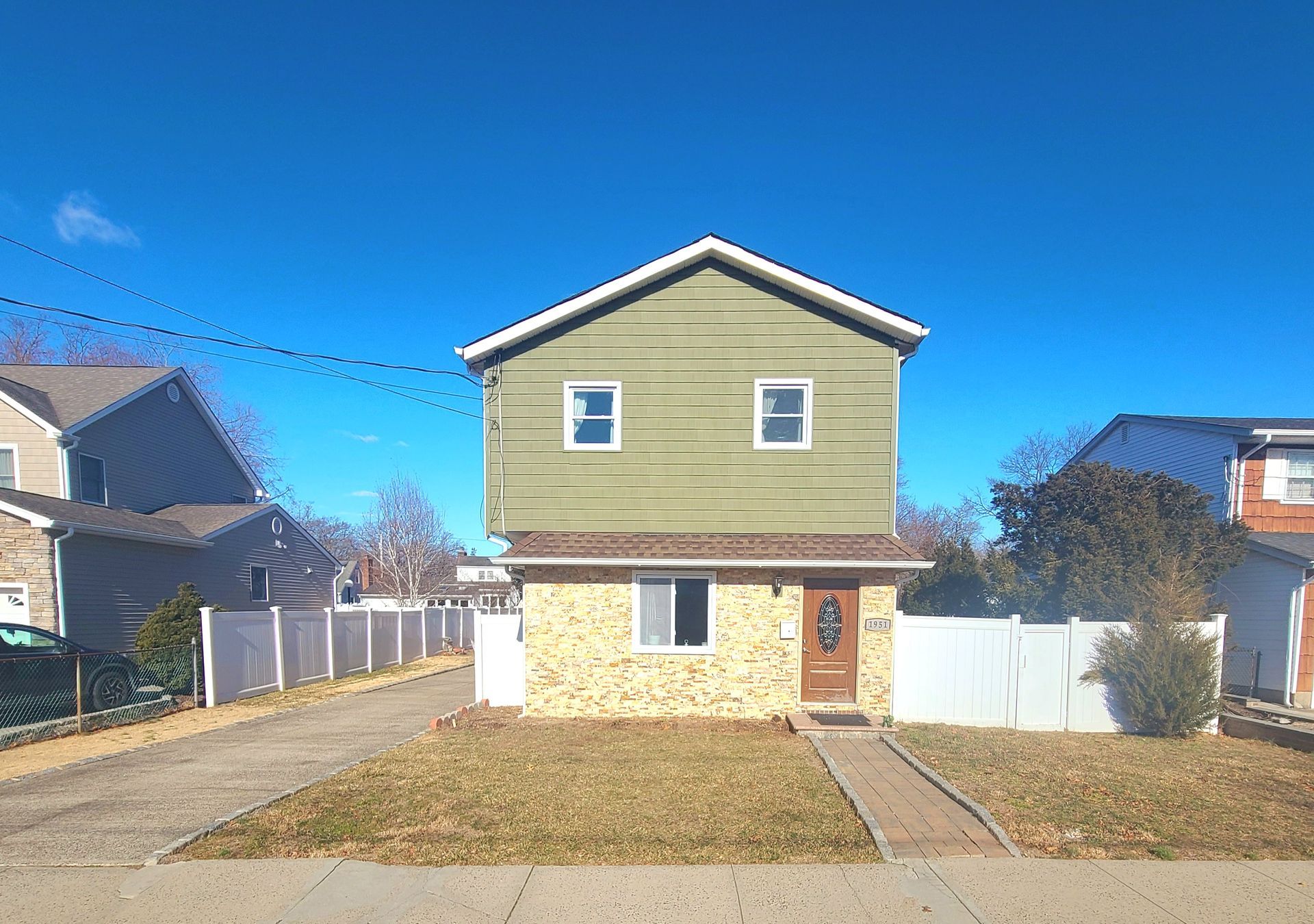 Two-story house with green siding on top, brick below, blue sky, white fence, and driveway in front.