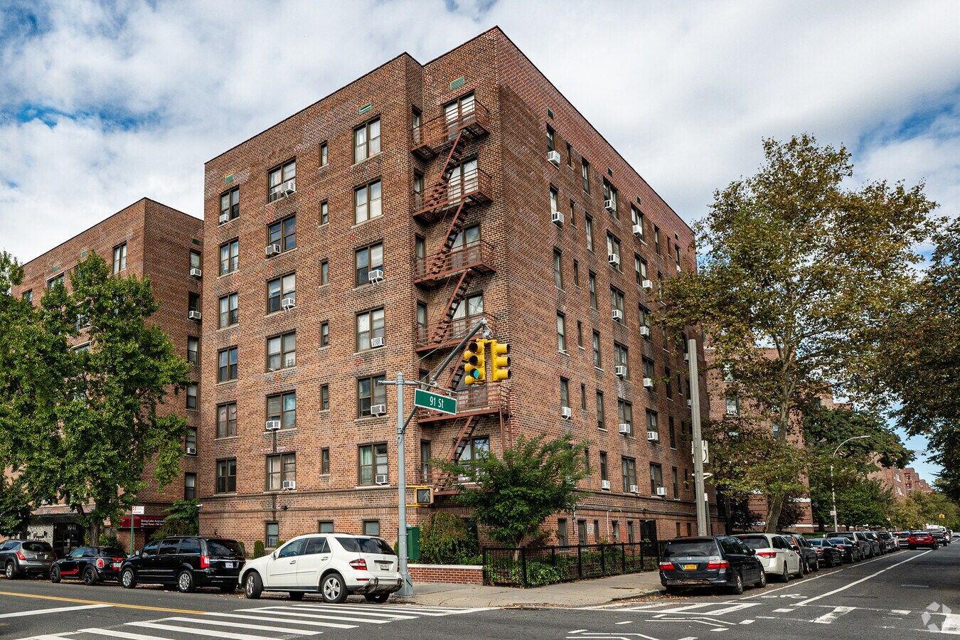Brick apartment building with fire escapes, street, parked cars, and trees under a cloudy sky.