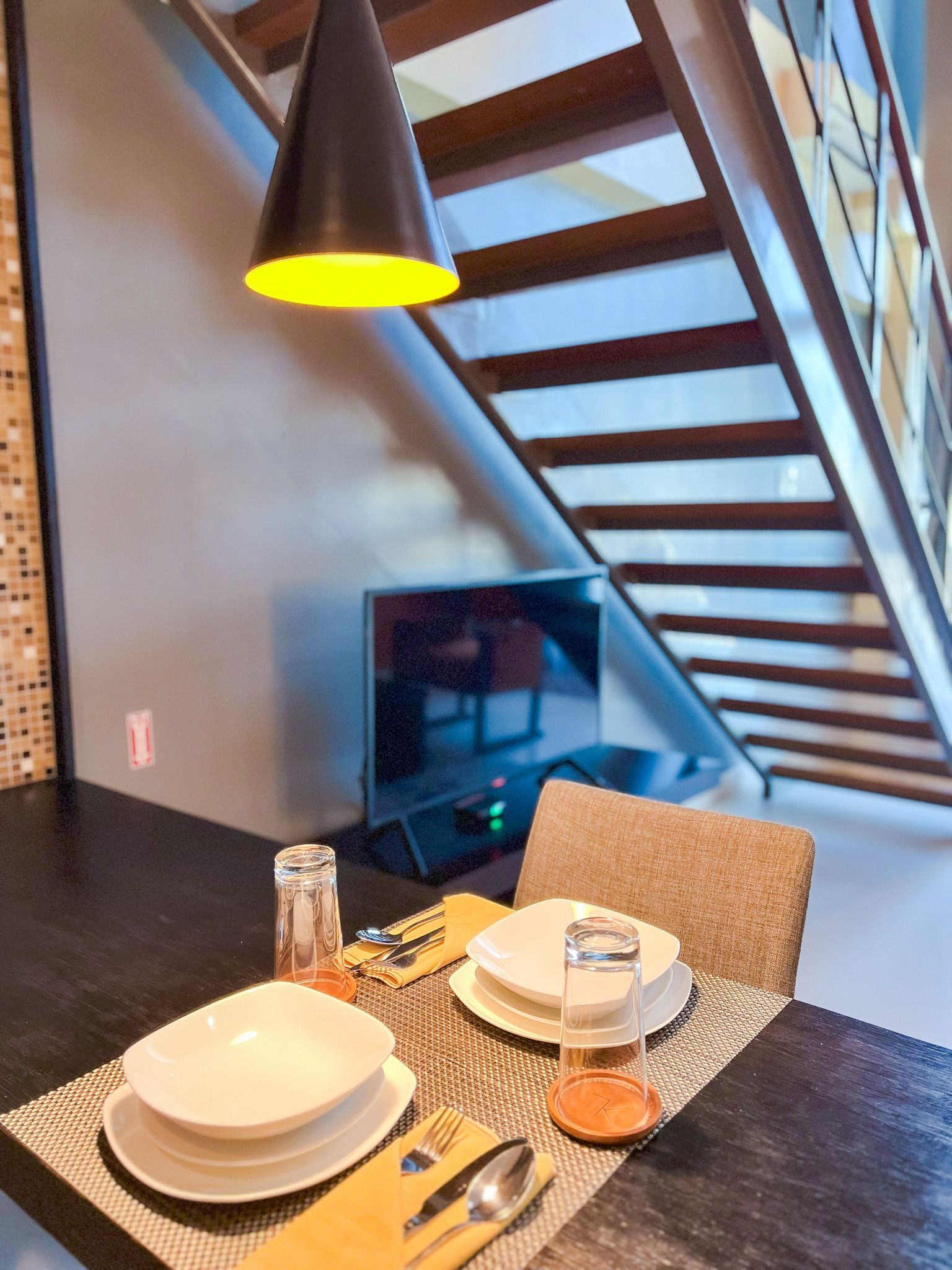 Dining area with table set for one, under a stairwell with a hanging light and TV.