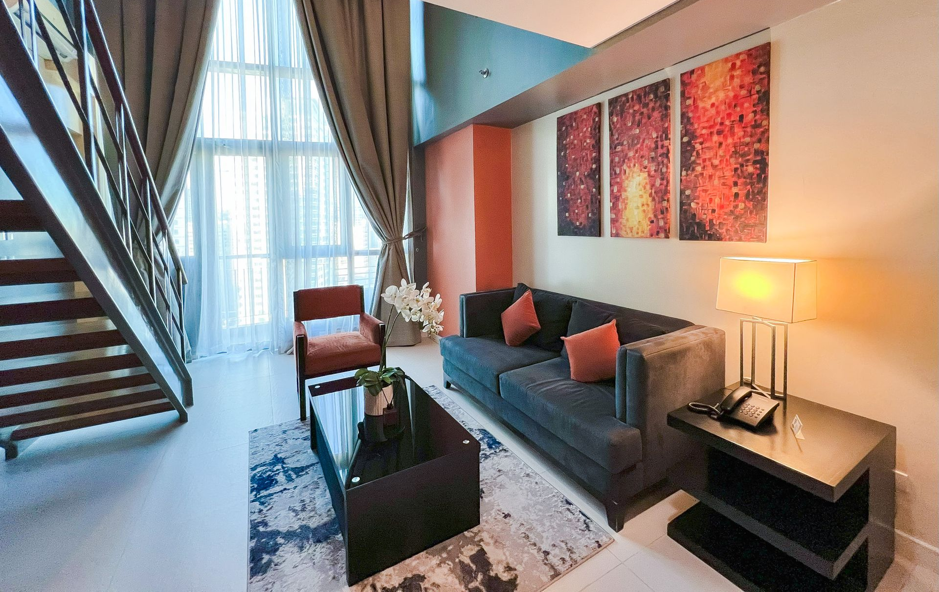 A modern living room with a dark sofa, coffee table, orange armchair, and abstract art on a wall by a staircase.