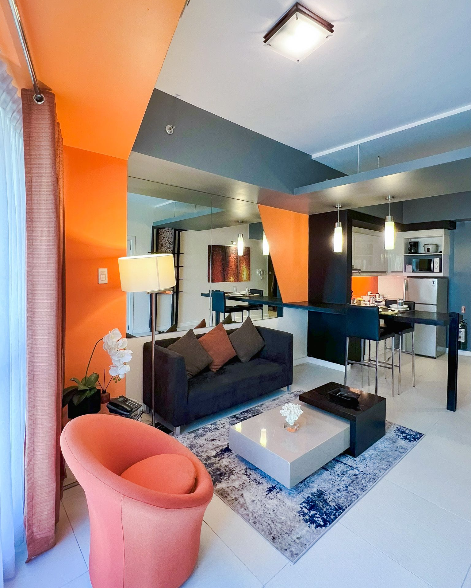 Living room with orange and gray walls, dark sofa, coral chair, kitchen in the background.