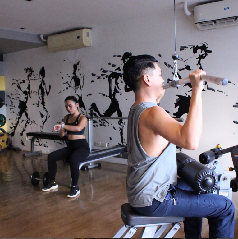 Man using lat pulldown machine in a gym. Woman in the background checks a smartwatch.