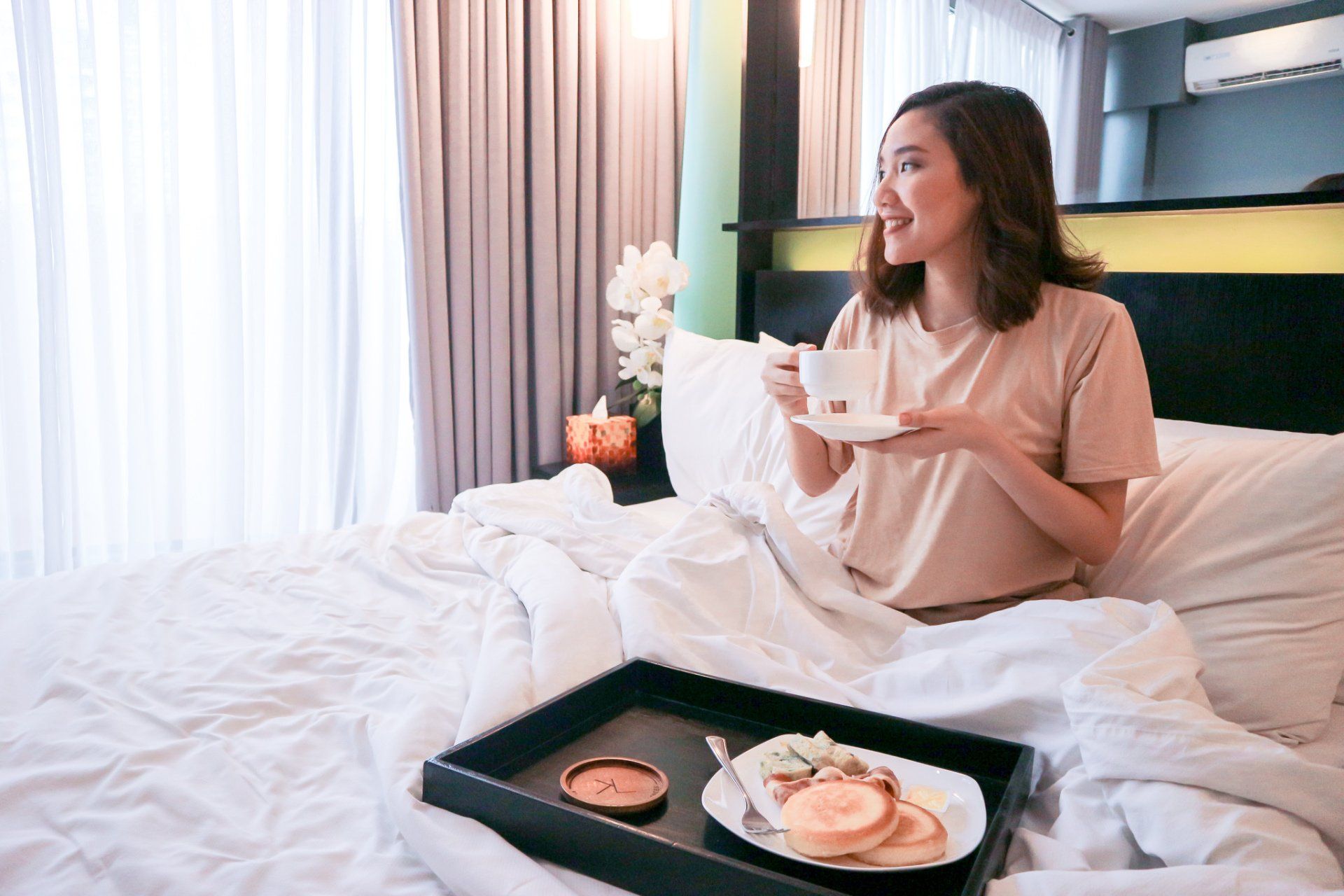 Woman in bed, smiling, holding a cup. Breakfast tray with pancakes and a dessert. Soft lighting.