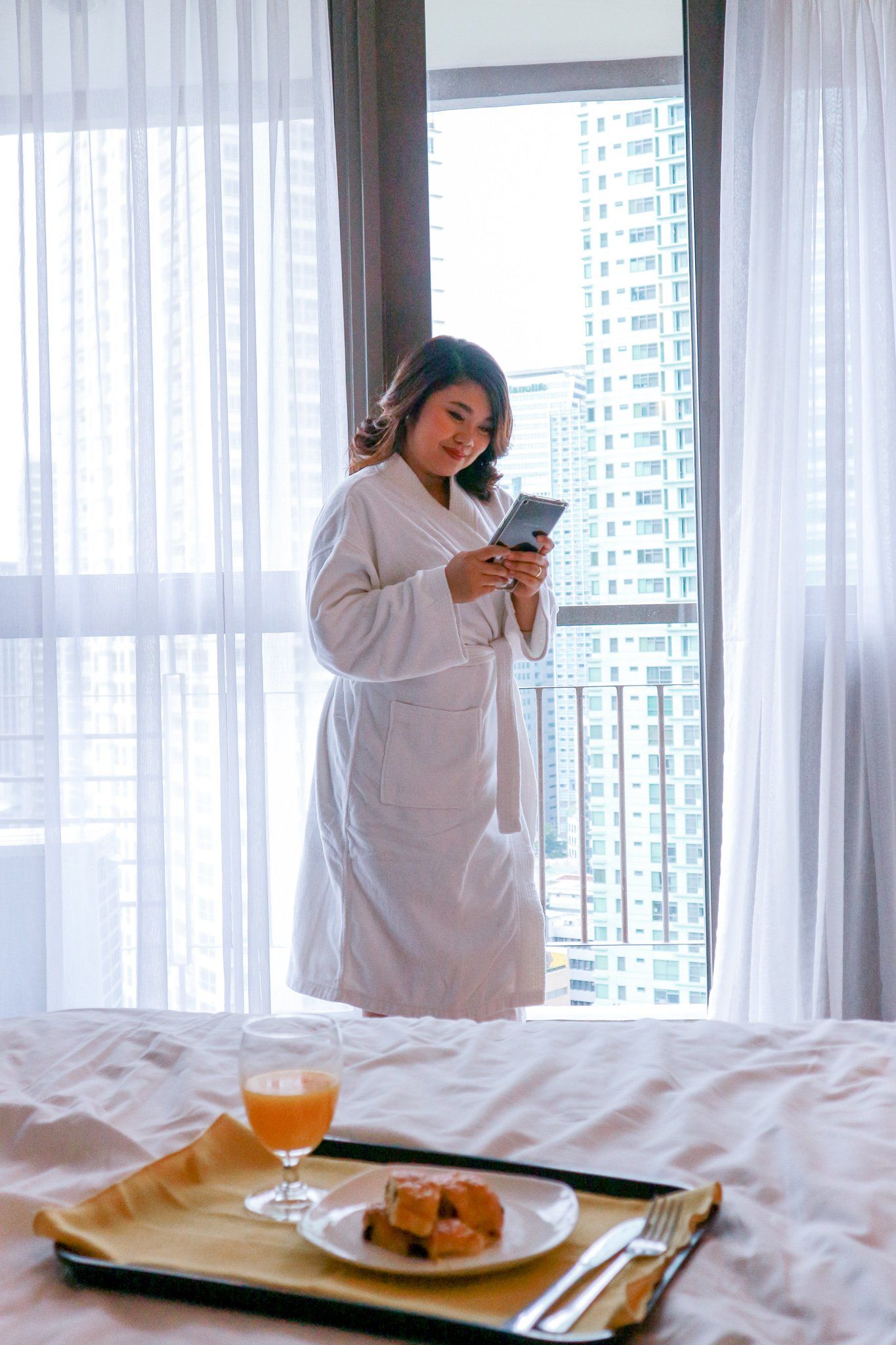 A woman in a bathrobe is standing next to a tray of food on a bed in a Makati hotel with pool