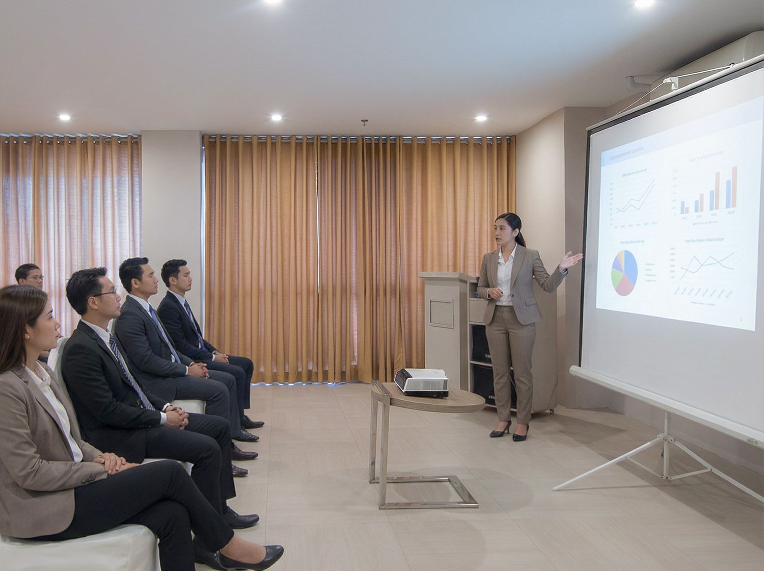 A presenter stands before a screen showing charts while a group in business attire watches in a conference room.