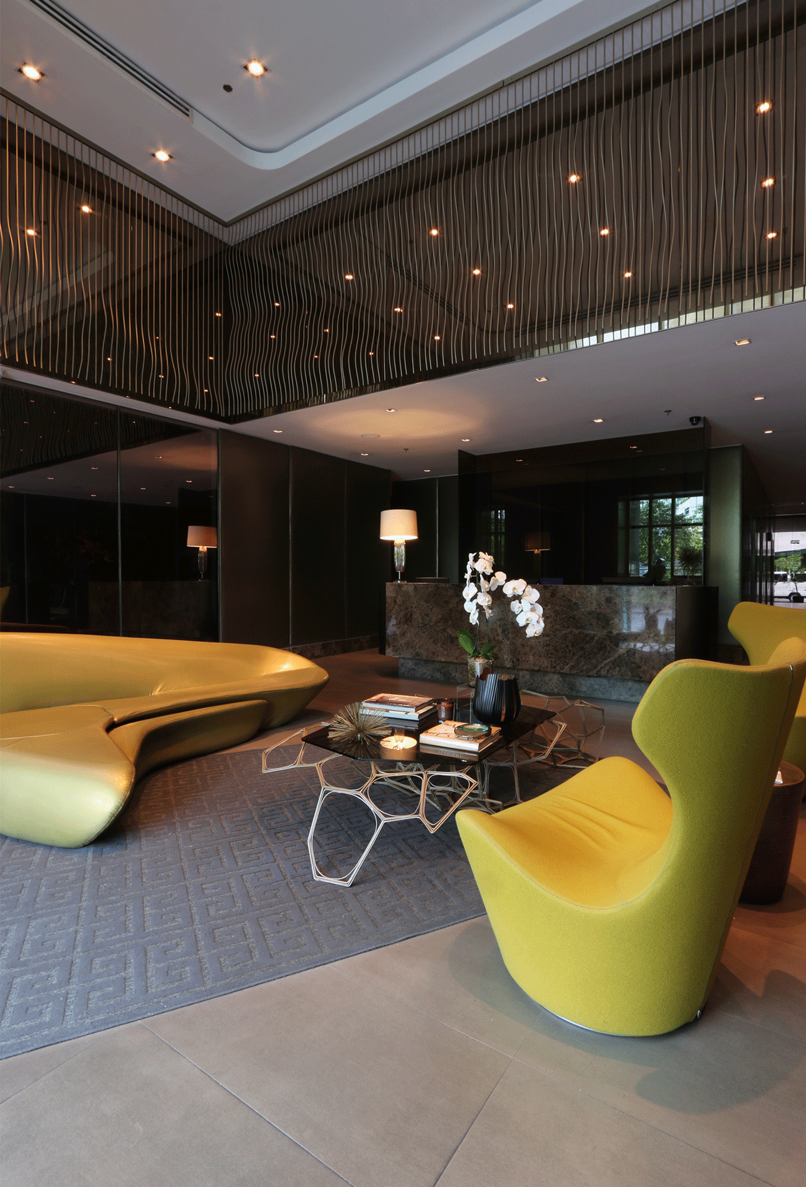 Modern hotel lobby with gold couch, yellow chair, patterned rug, and a front desk.