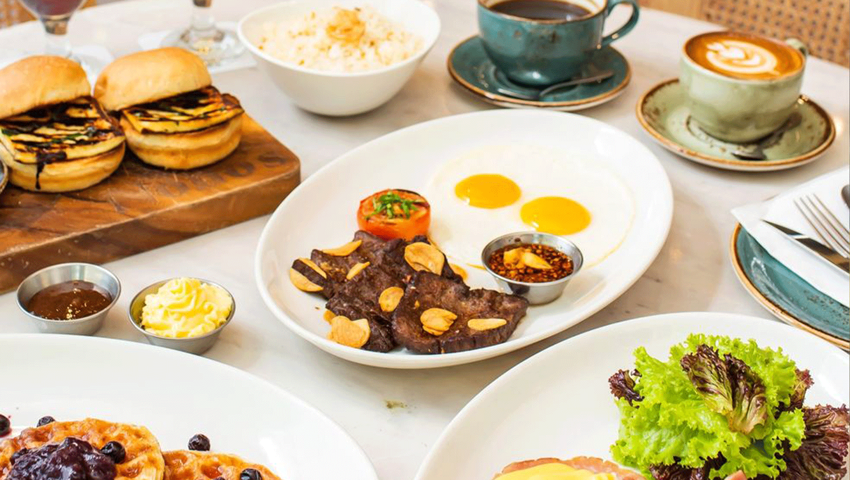 A breakfast spread on a table with sliders, garlic steak and eggs, waffles, salads, and two mugs of coffee.
