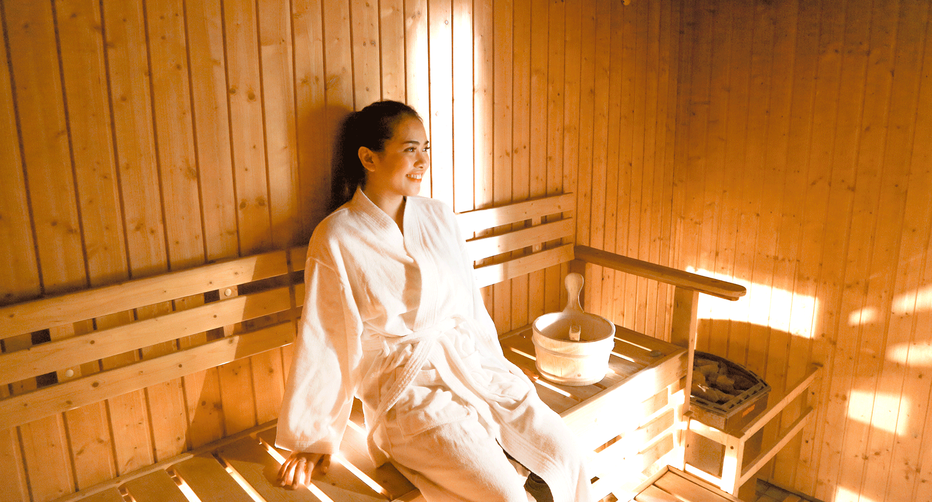 A person in a white robe relaxing in a wood-paneled sauna with a bucket on the bench.