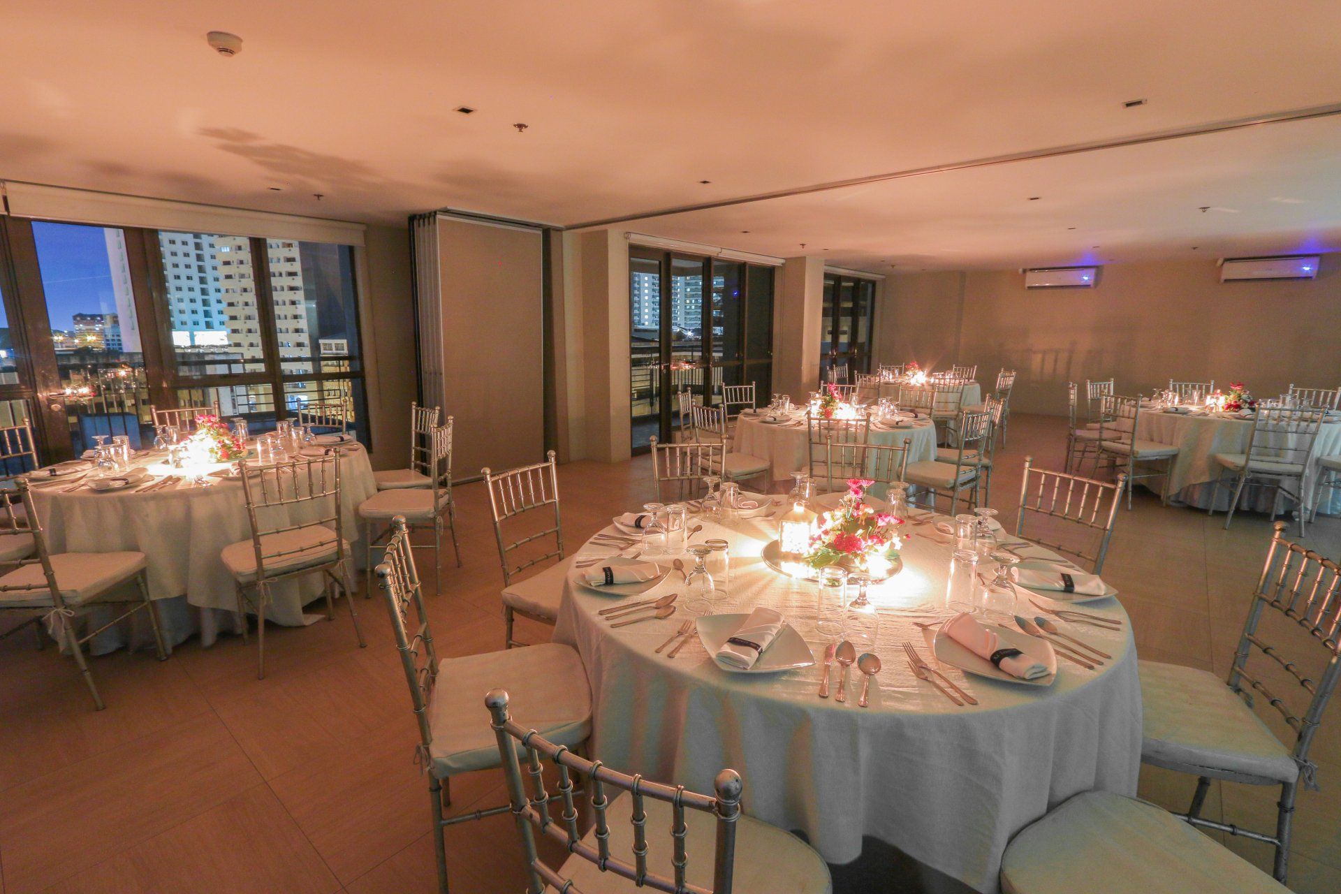 Elegant dining room set for a celebration at night. Tables are set with candles, flowers, and white linens. City lights visible outside.