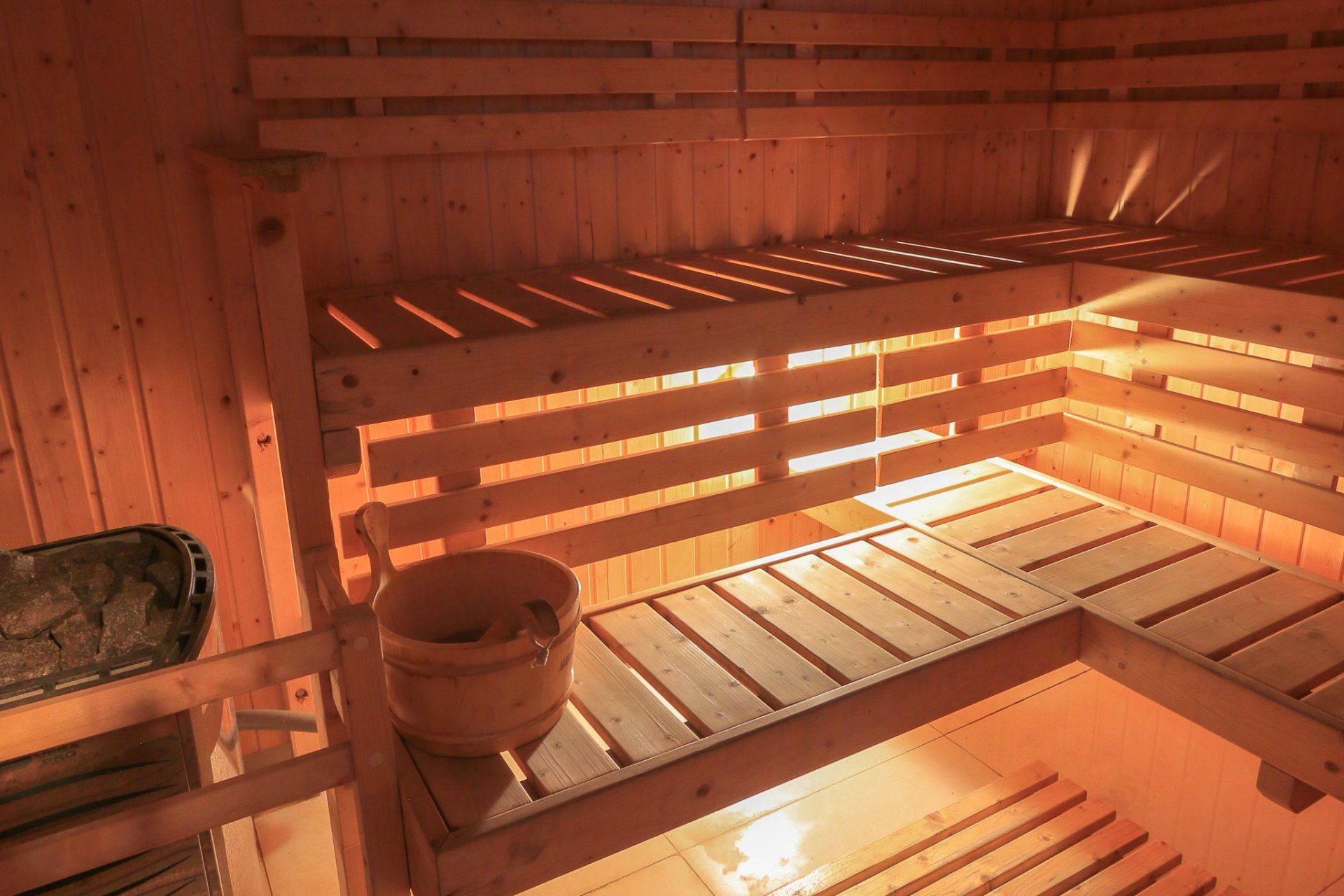 Wooden sauna interior with benches, bucket, and heating stones. Warm, glowing light.