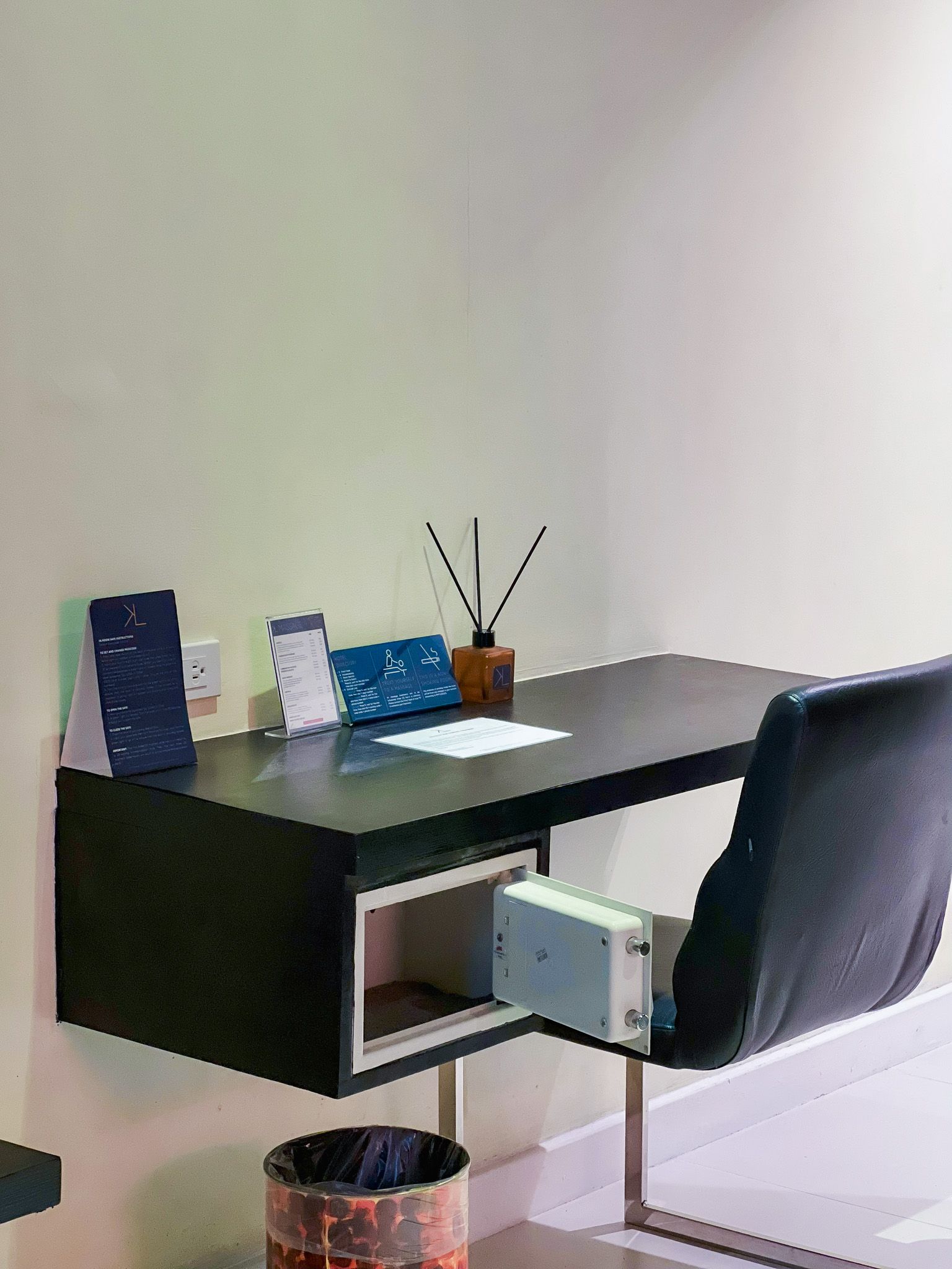 Black desk with a chair, safe, and trash can. Diffuser and papers sit on the desk.