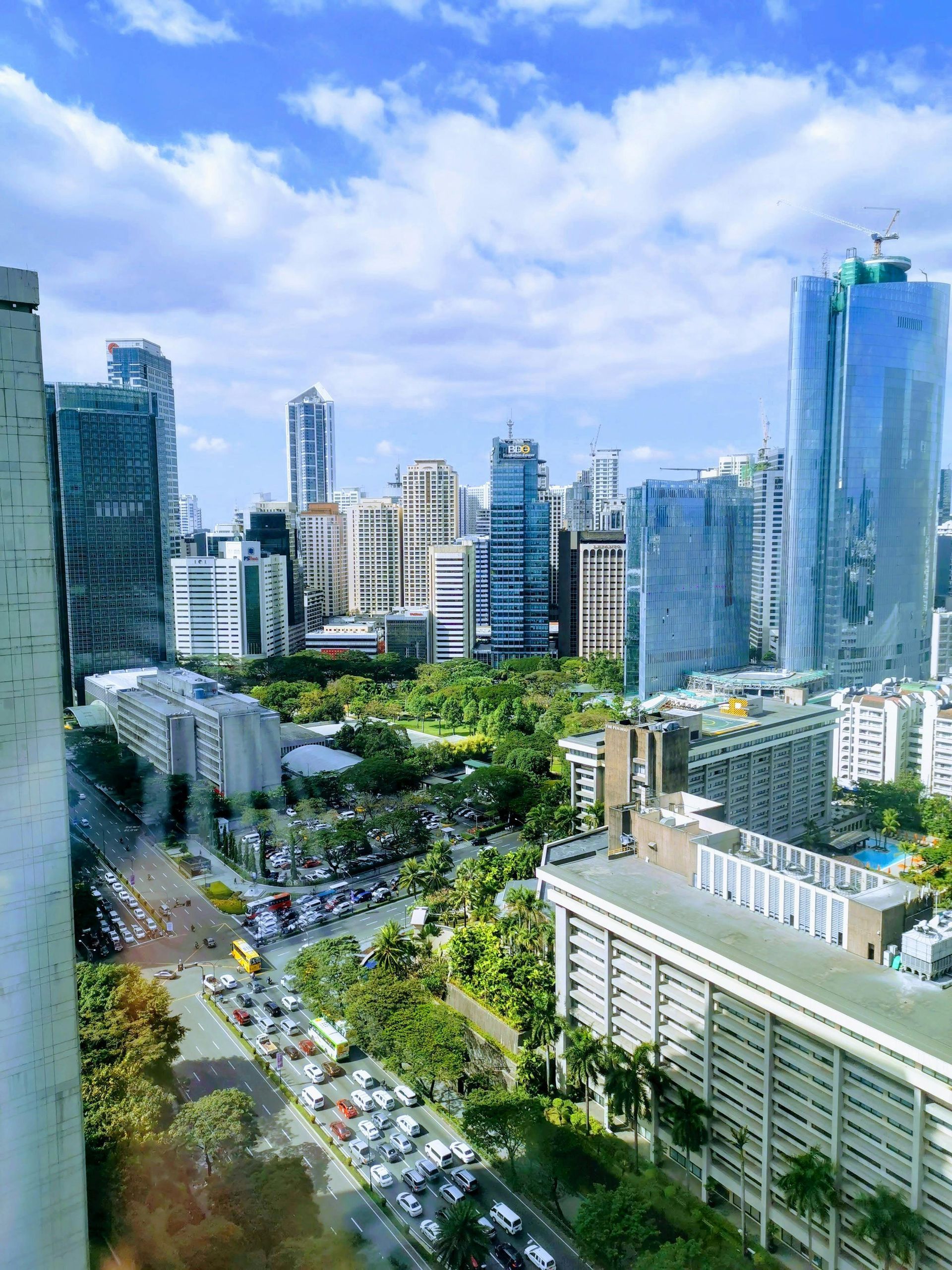 City skyline with tall glass buildings, a tree-lined street, and traffic under a bright blue sky.