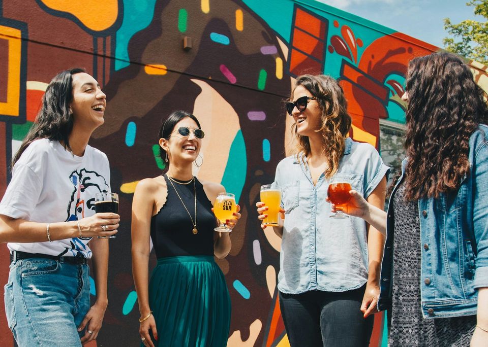 A group of women are standing in front of a colorful wall holding drinks.
