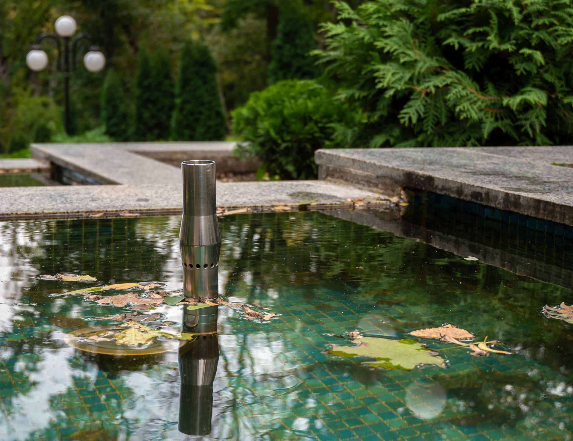 Fountain with stainless steel nozzle, concrete pool, and trees in background.