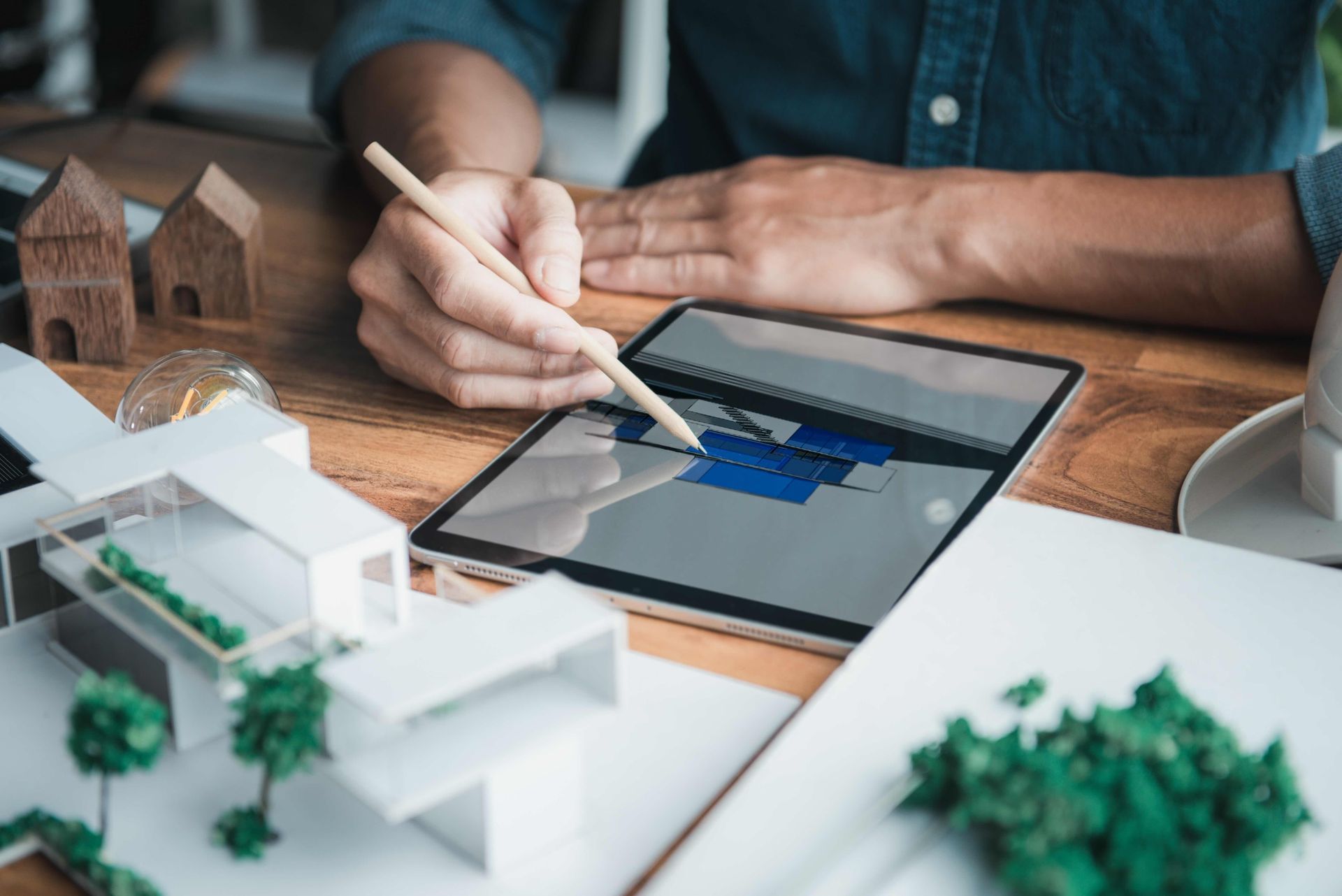 Architect using a stylus on a tablet, working on a building design. Models, trees, and tools on a wooden desk.