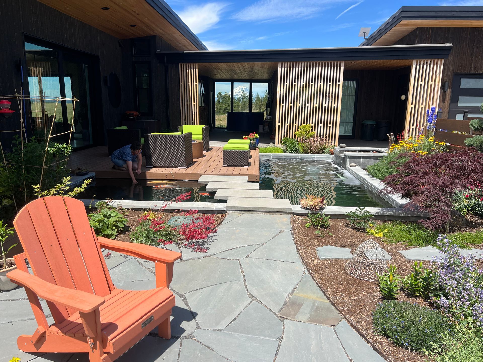 Orange Adirondack chair on a stone path, leading to a patio with a pond and seating area. Sunny day.