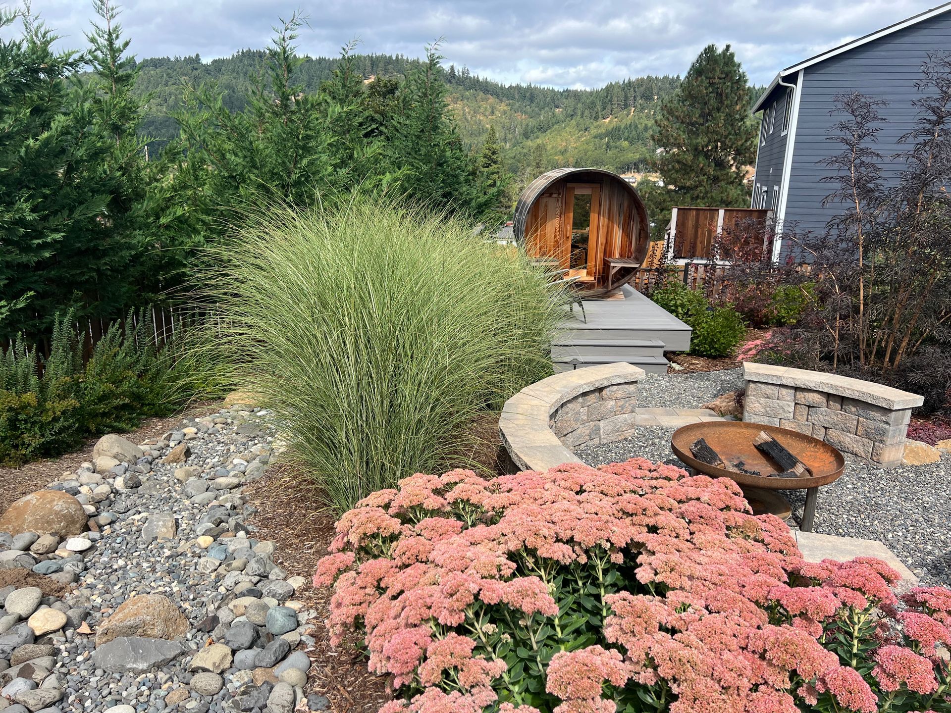 Backyard with hot tub, fire pit, and colorful landscaping, including pink flowers and a wooded mountain backdrop.