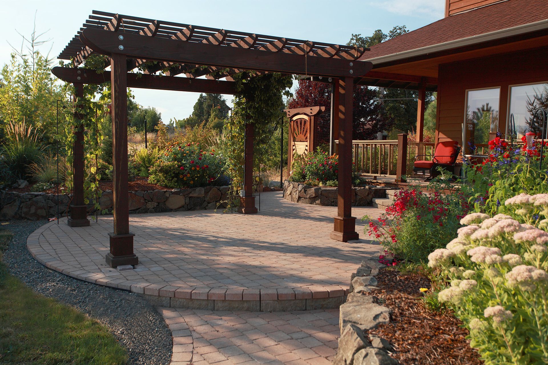 Pergola over a brick patio, surrounded by garden beds and a house.