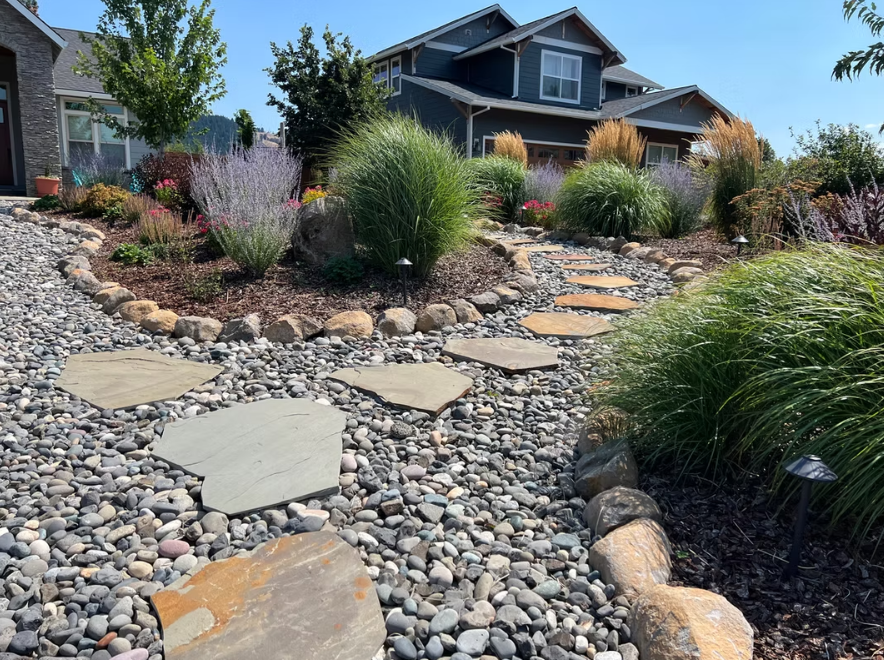 Stone path through a colorful garden bed in front of a two-story house with blue siding.