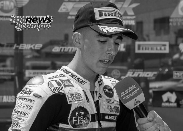 Motorcycle racer in white racing suit with cap, being interviewed; holding microphone.