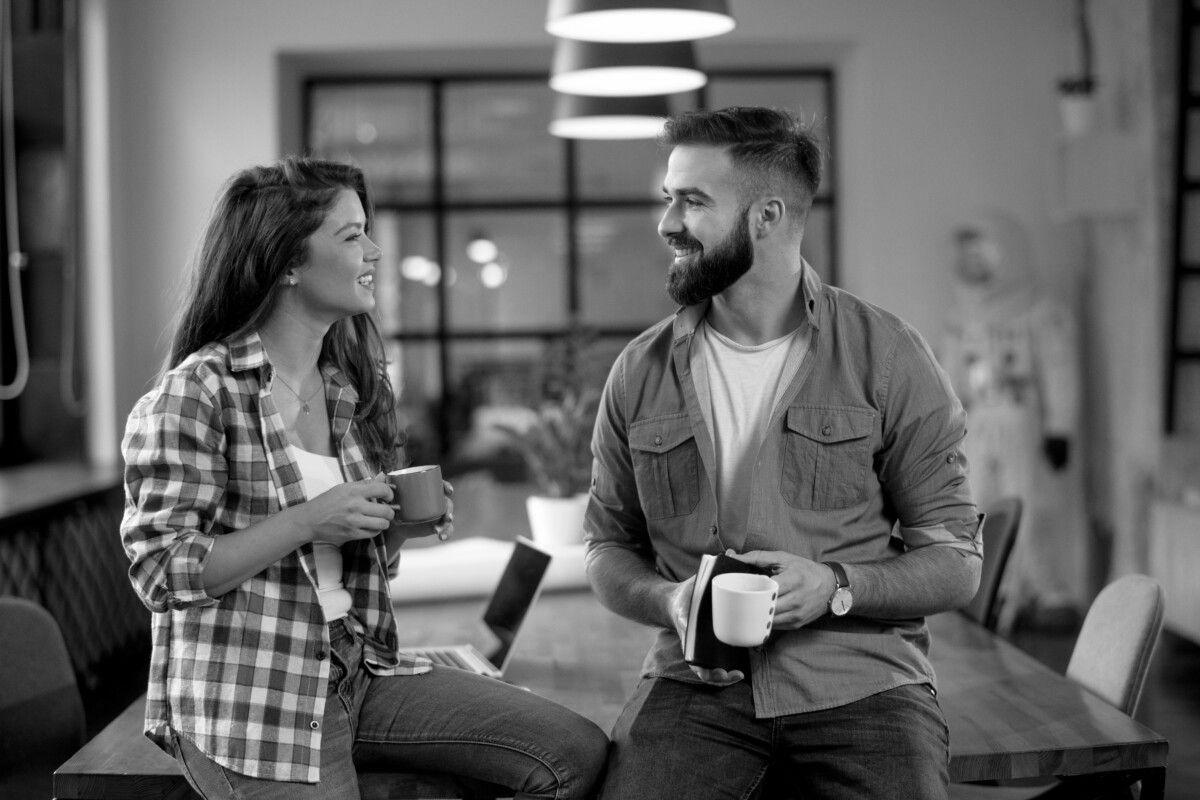 Woman and man with coffee cups converse, leaning on a table in an office setting.