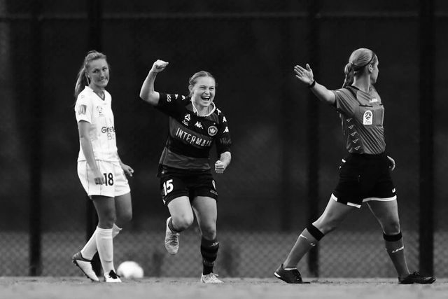 Soccer player celebrates a goal with raised arm, referee signals it good, another player looks on.