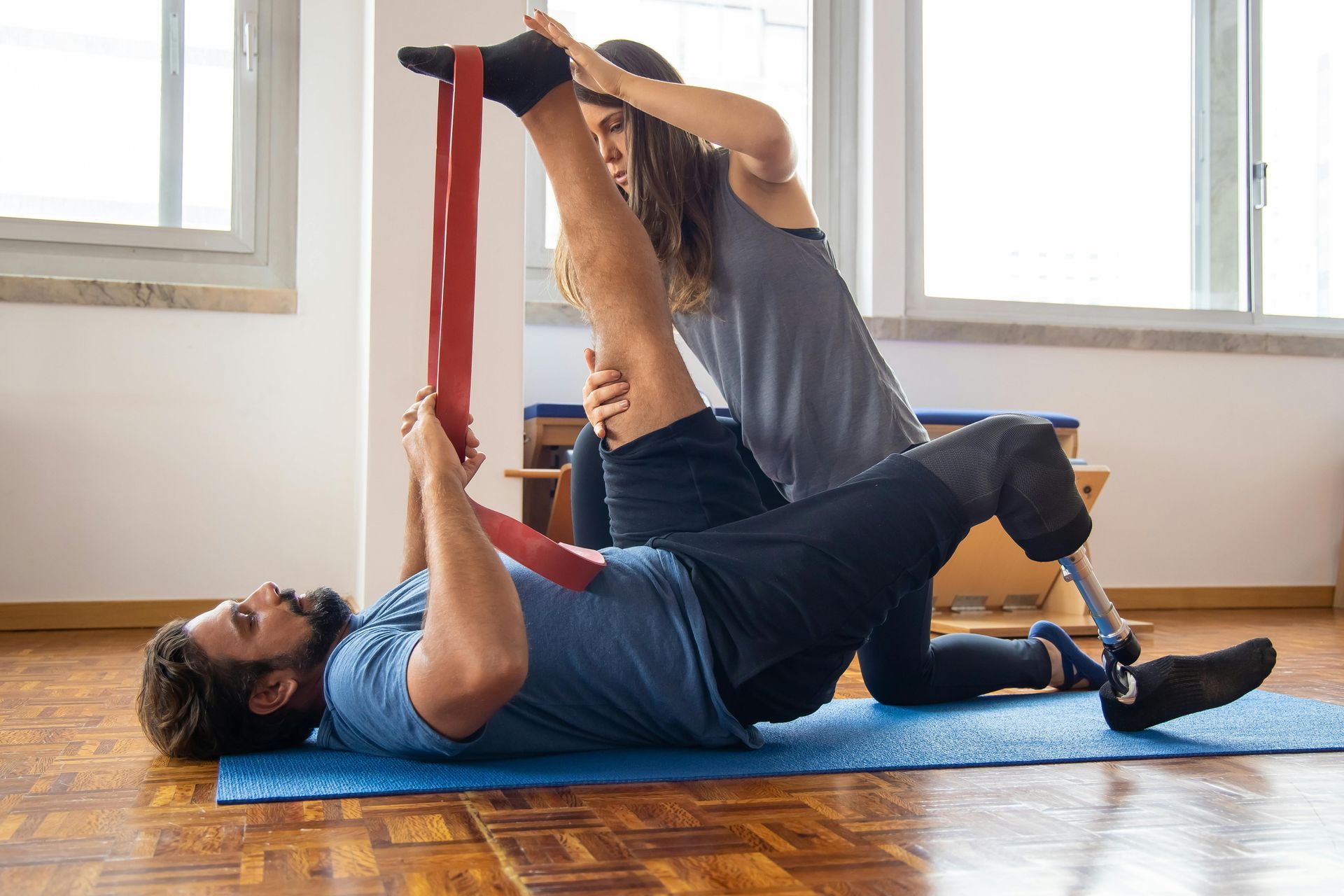 Man with prosthetic leg stretching with help of woman using a resistance band on blue mat.