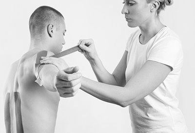 A person receives kinesiology tape on their shoulder by a medical professional in a white-walled room.