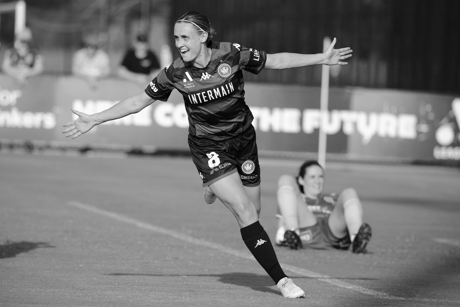 Soccer player in black jersey celebrates a goal with arms outstretched on a field; another player on the ground.